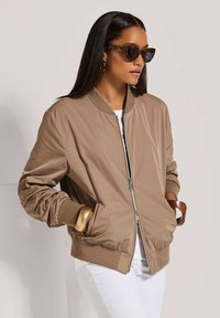 Vrouw met lang donker haar draagt een bril met schildpadmotief, een beige bomberjack, witte broek en forse gouden en houten armbanden, staand met haar handen in de zakken.