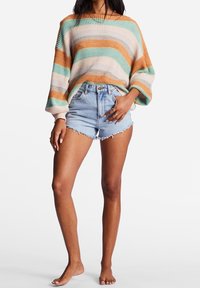 Übergroßer, gestrickter Pullover mit horizontalen Streifen in Orange, Grün und Weiß. Kombiniert mit zerrissenen, hochgeschnittenen Denim-Shorts.