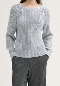 Maglione a coste grigio chiaro con scollatura rotonda e maniche lunghe, caratterizzato da una superficie texturizzata e orlo svasato. Indossato con pantaloni scuri.