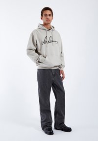Sudadera color beige claro con impresión negra de firma, bolsillo tipo canguro y capucha con cordón. Combinada con jeans holgados de color gris oscuro y zapatos negros.