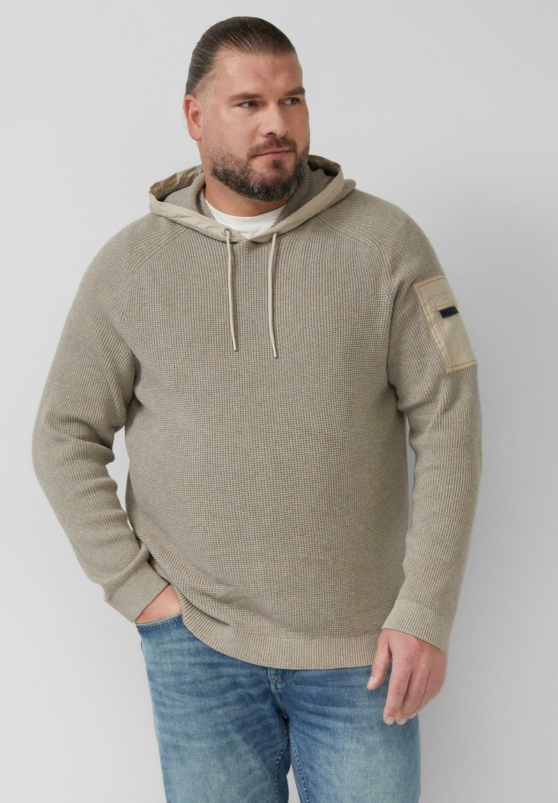 Beige gewatteerde hoodie met een trekkoord bij de hals, raglan mouwen en een zakaccent op de linkermouw. G gedragen met blauwe jeans.