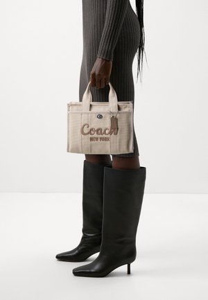 Sac fourre-tout en toile beige avec "Coach" et "New York" brodés en marron, doté de poignées courtes et d'une étiquette en cuir. Le modèle porte des bottes noires jusqu'au genou.