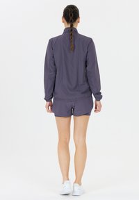 Lilla træningsjakke med høj krave, lange ærmer og elastiske manchetter, parret med matchende shorts og båret med hvide sneakers.