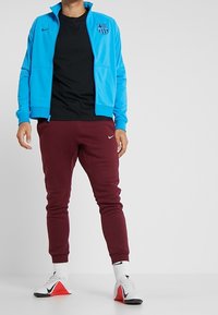 Mann in einer leuchtend blauen FC Barcelona-Jacke, schwarzem Hemd, kastanienbraunen Nike-Sweatpants, weißen Socken und weißen Nike-Sneakern mit roten Sohlen.