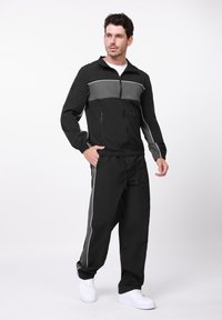 Μαύρη αθλητική φόρμα από ελαφρύ υλικό με γκρι λεπτομέρειες, που περιλαμβάνει μπουφάν με φερμουάρ και παντελόνι με ρυθμιζόμενη μέση και πλάγιες ρίγες.