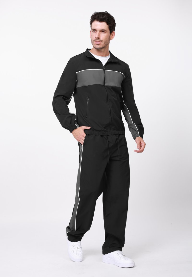 Μαύρη αθλητική φόρμα από ελαφρύ υλικό με γκρι λεπτομέρειες, που περιλαμβάνει μπουφάν με φερμουάρ και παντελόνι με ρυθμιζόμενη μέση και πλάγιες ρίγες.