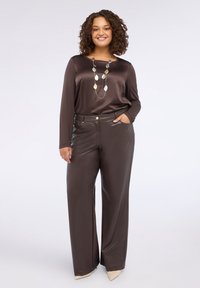 Top lungo in satin marrone a maniche lunghe abbinato a pantaloni larghi lucidi marroni. Accessoriato con una collana con pendente multimetallico e tacchi beige.