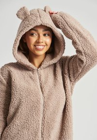 Fuzzy, lynlås hættebluse i lys beige med bjørneører. Blødt, tekstureret materiale, lange ærmer og en afslappet pasform.