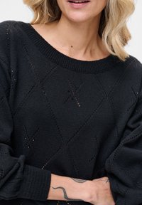 Czarny dziergany sweter z wzorem diamentowym, ozdobiony małymi oczkami, z szerokim dekoltem i elastycznymi mankietami, nadający fakturowany wygląd.