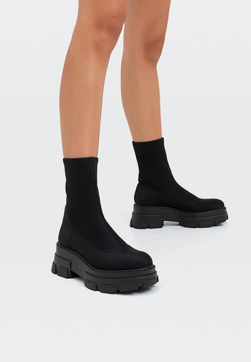 Bottes noires à la cheville en tissu extensible avec une semelle plateforme épaisse. Texture lisse, tige haute et design minimaliste.