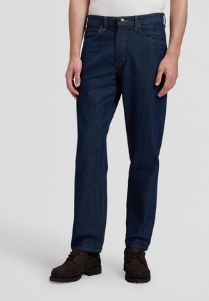 Mann trägt dunkelblaue Jeans mit geradem Bein und dunkelbraune Schnürschuhe, steht vor einem schlichten hellen Hintergrund.