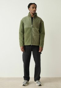 Grøn fleecejakke med lynlås foran, der har tekstureret stof på den nederste halvdel og blødt corduroy på den øverste, båret over sorte cargobukser.