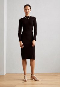 Musta ribbed-neulemekko, jossa on pitkät hihat, tiukka siluetti ja polvimittainen helma. Yhdistetty vaaleanbeigeihin sandaaleihin, joissa on matala lohko korko.