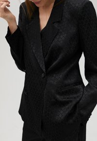 Černý blazer vyrobený z texturované tkaniny s jemným geometrickým vzorem, s jedním knoflíkem a dvěma předními kapsami.