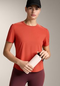 Piros sportos rövid ujjú póló kerek nyakkivágással, egy világos rózsaszín vízpalackkal, fekete kupakkal a tetején tartva.