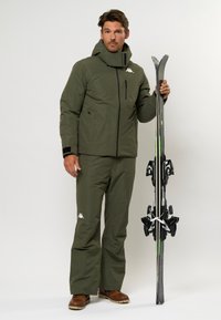 Zielona wodoodporna kurtka narciarska z wysokim kołnierzem i kieszenią na zamek, zestawiona z dopasowanymi narciarskimi spodniami. Model trzyma czarne narty z wiązaniami.