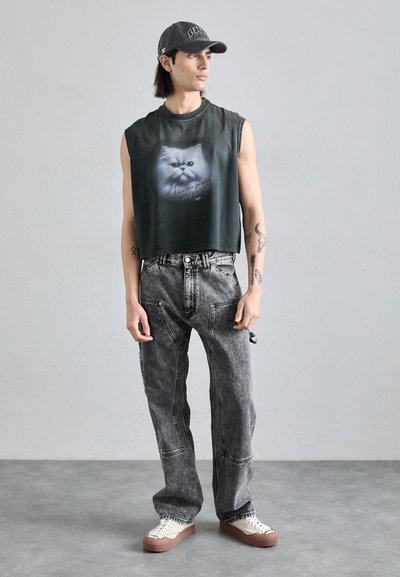 Black sleeveless cropped t-shirt with a cat graphic. Gray loose-fit jeans with side pockets. Cream sneakers with brown soles. Gray background.