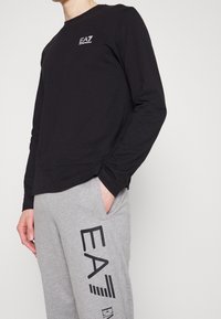 Černé triko s dlouhým rukávem a logem EA7, doplněné šedými tepl áky s velkým černým potiskem EA a detaily bočních kapes.