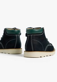 Des bottes en daim bleu marine présentent un col en cuir vert, des coutures blanches contrastantes et une semelle en caoutchouc avec un design de grip texturé.