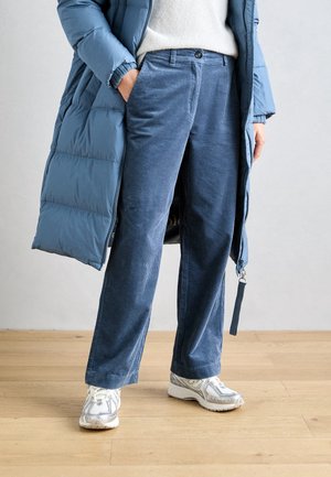 Person iført blå kordfløjlsbukser, hvide sneakers, en hvid sweater og en lang blå polstret jakke, stående på et trægulv.