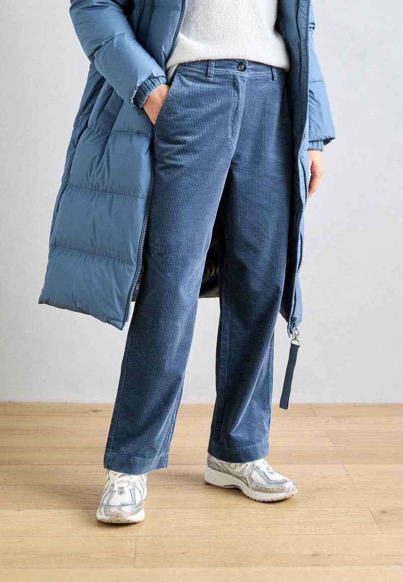 Personne portant un pantalon en velours côtelé bleu, des baskets blanches, un pull blanc et un long manteau matelassé bleu, debout sur un sol en bois.