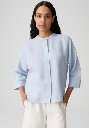 Frau mit langen dunklen Haaren, trägt ein hellblaues Hemd mit 3/4-Ärmeln und weiße Hose, Hände in den Taschen, neutraler Hintergrund.