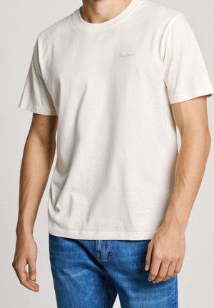 Cremefarbenes Baumwoll-T-Shirt mit kurzen Ärmeln, rundem Ausschnitt und kleinem Logo oben links. Kombiniert mit blauen Jeans.