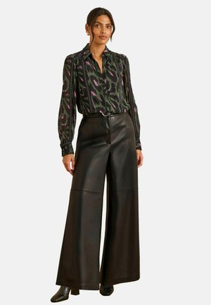 Schwarze, lederne weit geschnittene Hosen kombiniert mit einer Bluse in Grün, Lila und Schwarz mit Muster, die einen Kragen und Knopfdetails aufweist.