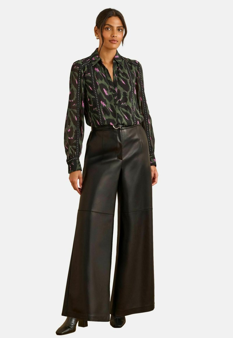 Melni ādas platās bikses, kas apvienotas ar zaļu, purpura un melnu rakstainu blūzi ar apkakli un pogu detaļām.