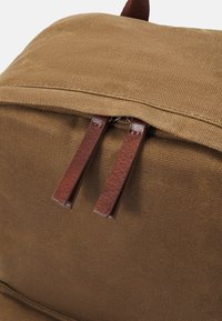 Hnědý plátěný batoh s horní klopou, s hnědými koženými táhly na zip a texturovanou látkou. Pevná konstrukce s odolnými materiály.