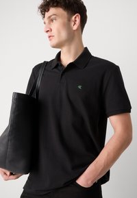 Zwarte polo in een gebreide stof, met een groen logo op de borst. Vergezeld van een zwarte leren tas met een zachte textuur.