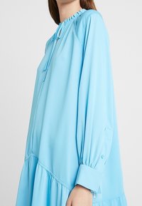 Ljusblå klänning i slät väv, med dragsko i halsen, långa ärmar med knappmanchetter och en lager-p å-lager nederdel.