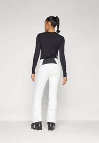 Zwarte longsleeve shirt met een getailleerd ontwerp, gecombineerd met witte skibroeken met een zwart accent op de achterkant. Zwarte skischoenen zichtbaar.