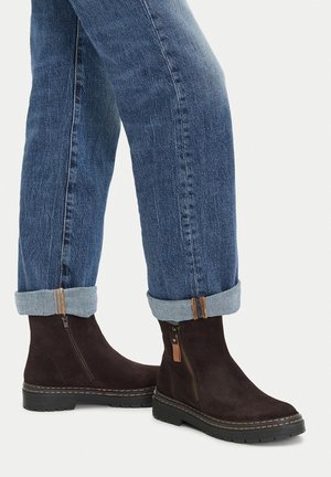 Brązowe zamszowe botki z bocznym zamkiem i brązowym skórzanym akcentem, zestawione z podwiniętymi jeansami. Solidna czarna gumowa podeszwa.