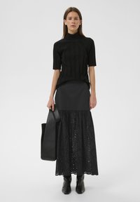 Schwarzer, gerippter, kurzärmeliger Rollkragenpullover kombiniert mit einem langen schwarzen Rock mit Ösenmuster, getragen mit einer großen schwarzen Tragetasche.