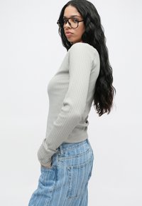 Γυναίκα με μακριά μαύρα κυματιστά μαλλιά, φορώντας μεγάλα γυαλιά, ανοιχτό γκρι ριμπ μανίκι μακρύ μπλούζα και ψηλοκάβαλο ανοιχτό μπλε τζιν με τα χέρια στις τσέπες.