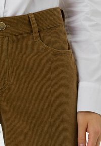 Brązowe spodnie sztruksowe z pionowym prążkowaniem, z zapięciem na guzik i bocznymi kieszeniami. W komplecie z białą koszulą z długim rękawem.