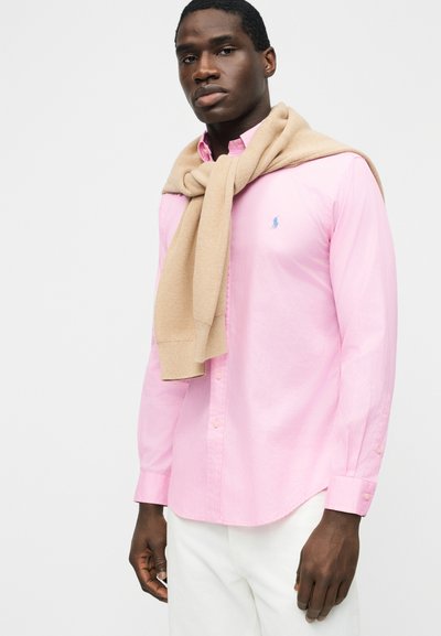 Muž oblečený v ružovej košeli na gombíky s béžovým svetríkom prehodeným cez ramená a bielymi nohavicami, stojaci na jednofarebnom pozadí.