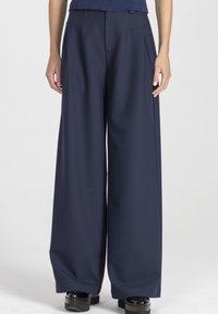 Szerokie spodnie w ciemnogranatowym materiale, z przodu zakładki, boczne kieszenie i gładka faktura. Zestawione z masywnymi czarnymi butami na platformie.