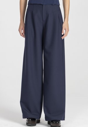 Szerokie spodnie w ciemnogranatowym materiale, z przodu zakładki, boczne kieszenie i gładka faktura. Zestawione z masywnymi czarnymi butami na platformie.