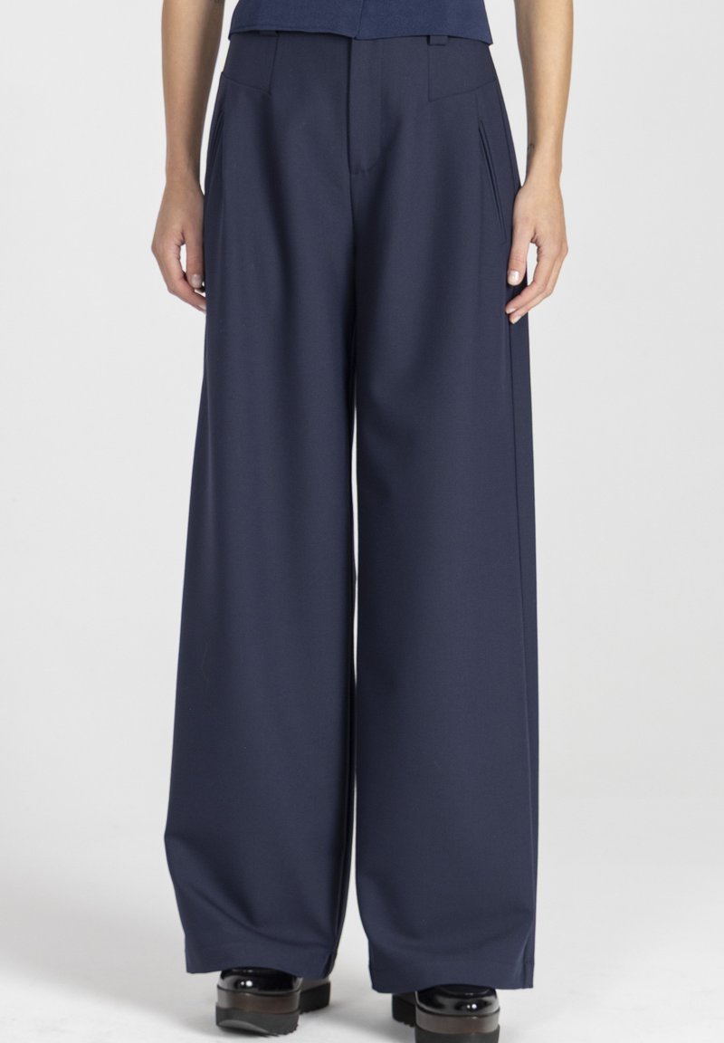 Szerokie spodnie w ciemnogranatowym materiale, z przodu zakładki, boczne kieszenie i gładka faktura. Zestawione z masywnymi czarnymi butami na platformie.