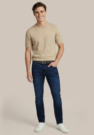 Jonge man glimlachend, gekleed in een beige overhemd met korte mouwen en zak, donkerblauwe jeans met gevlochten riem, en witte sneakers, staand tegen een effen achtergrond.