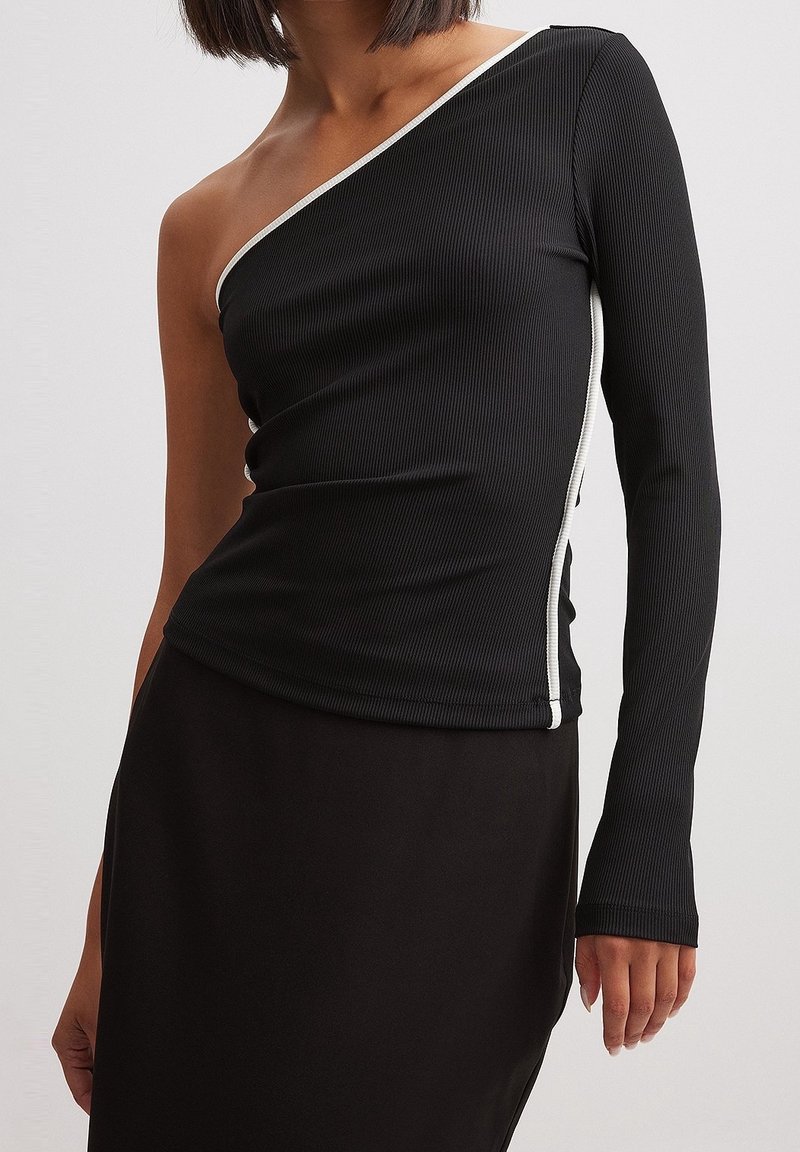 Vrouw draagt een zwart geribd een-schoudertop met één lange mouw en witte bies, gecombineerd met een zwarte rok.