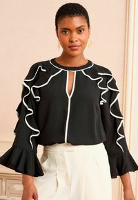 Blusa nera con accenti arricciati bianchi, scollatura a chiave e maniche lunghe. Il tessuto ha una texture liscia. Abbinata a pantaloni chiari.