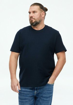 Man met baard en het haar naar achteren gebonden, gekleed in een donkerblauw T-shirt en blauwe spijkerbroek, staat met één hand in zijn zak en kijkt naar links.