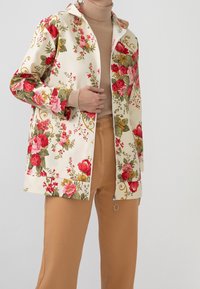 Persona vestida con un jersey de cuello alto beige, pantalones color camel y una chaqueta cremosa con cremallera decorada con un patrón floral en rojo, rosa, amarillo y hojas verdes.