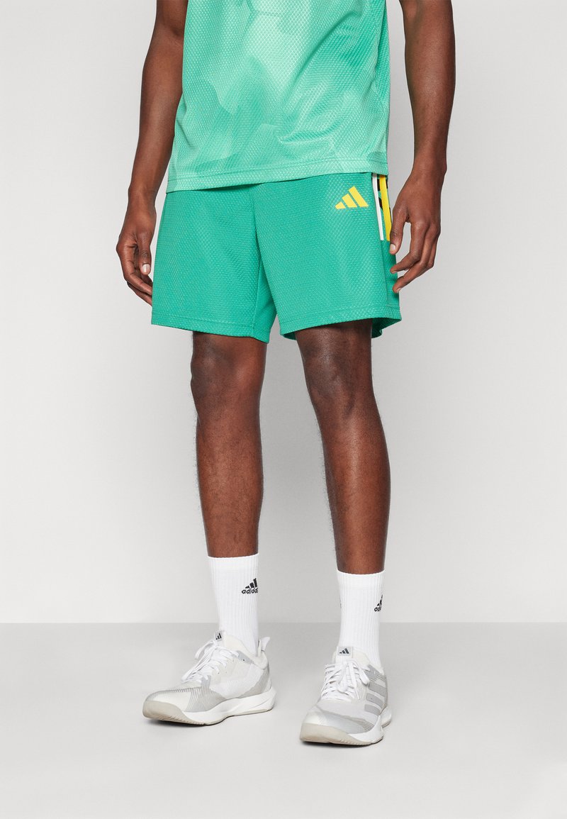 Green mesh athletic shorts with an elastic waistband, featuring a yellow Adidas logo and side pockets. Worn with white socks and gray shoes.