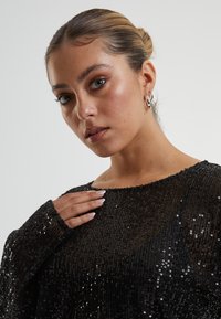 Crna sequinska majica s dugim rukavima i brodskim izrezom; ima teksturiranu površinu i slobodan kroj. Modelova ruka nježno se oslanja na tkaninu.