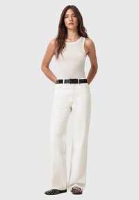 Pantaloni bianchi a gamba larga abbinati a una canotta beige chiaro. Presenta una cintura nera con borchie metalliche e scarpe nere slip-on con borchie decorativi.