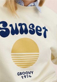 Kremowy sweatshirt z niebieskim tekstem "Sunset", żółtą grafiką słońca z poziomymi paskami oraz napisem "GROOVY 1976" na dole.